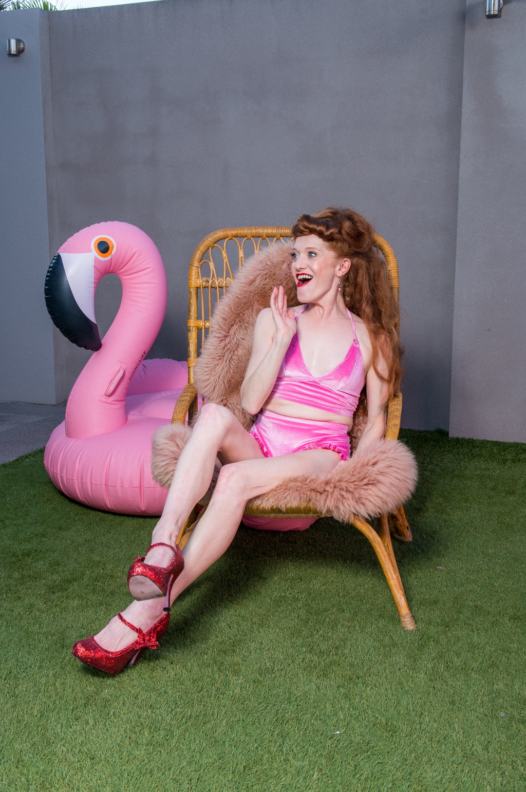 A person in a vibrant pink outfit and red heels sits on a wicker chair with a fluffy pink flamingo pool float nearby.