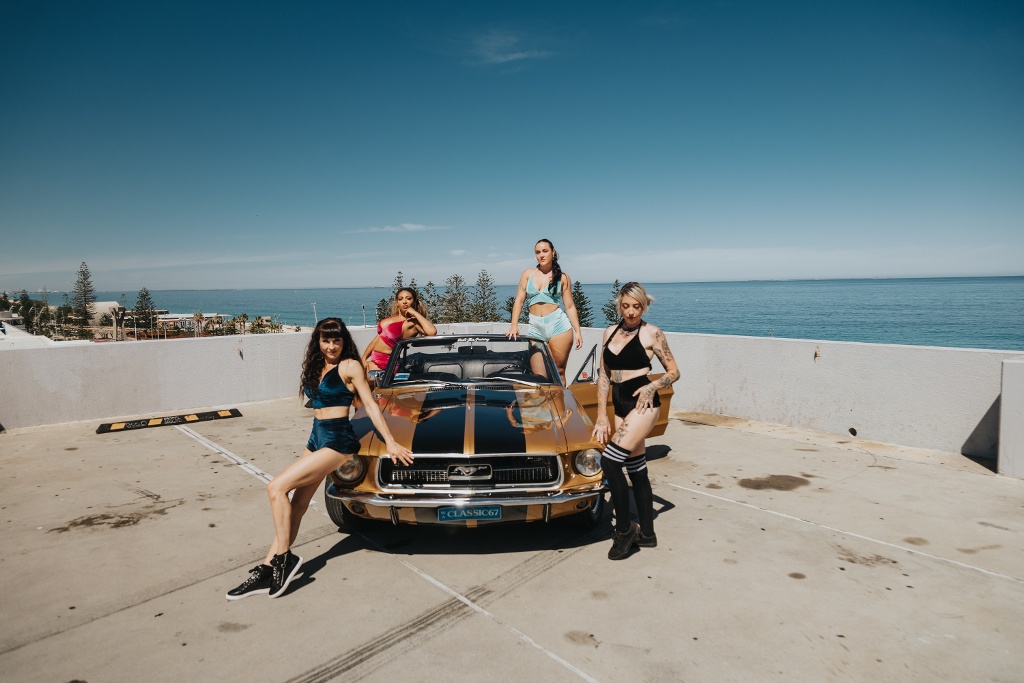 Four women posing confidently around a classic car on a rooftop overlooking the ocean.