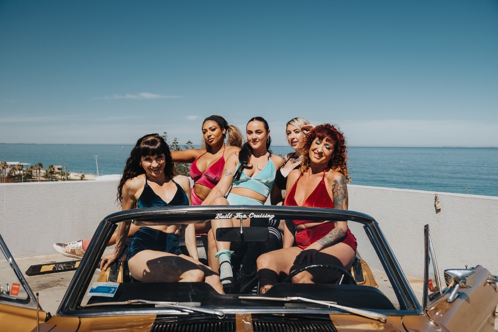 Five women posing together in a convertible car by the ocean on a sunny day.