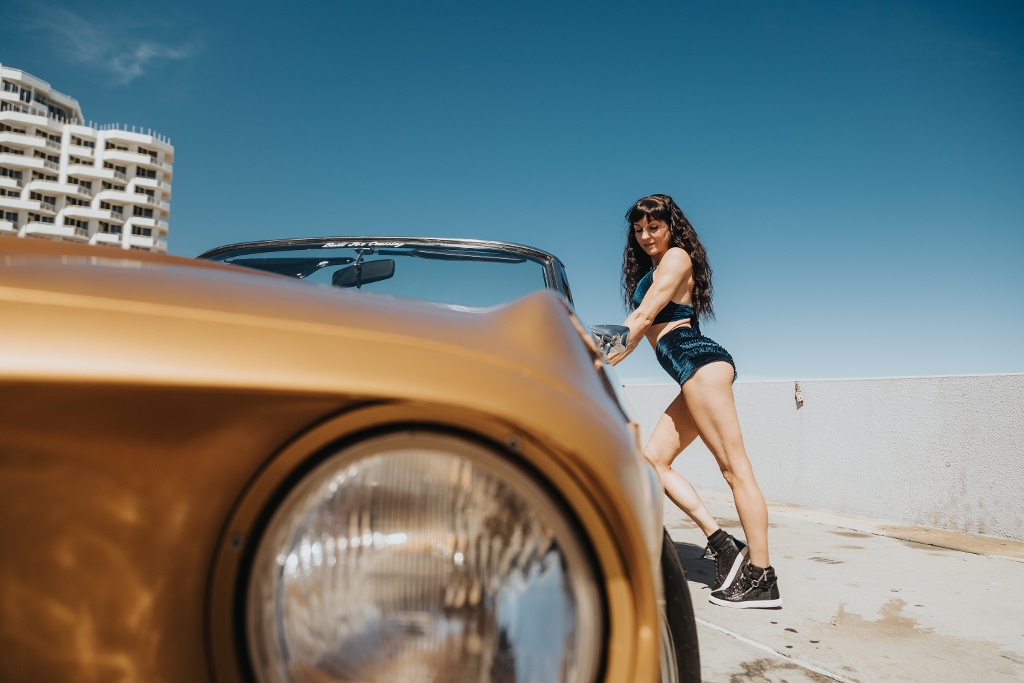 A person in a sporty outfit leans against a vintage car on a sunny day near a cityscape.