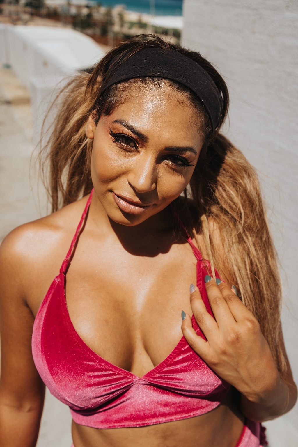 A person wearing a pink bikini top and a black headband poses outdoors with a sunny beach backdrop.