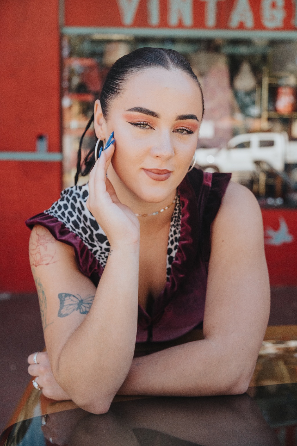 A person with bold makeup and colorful accessories poses confidently in front of a vintage-themed backdrop.