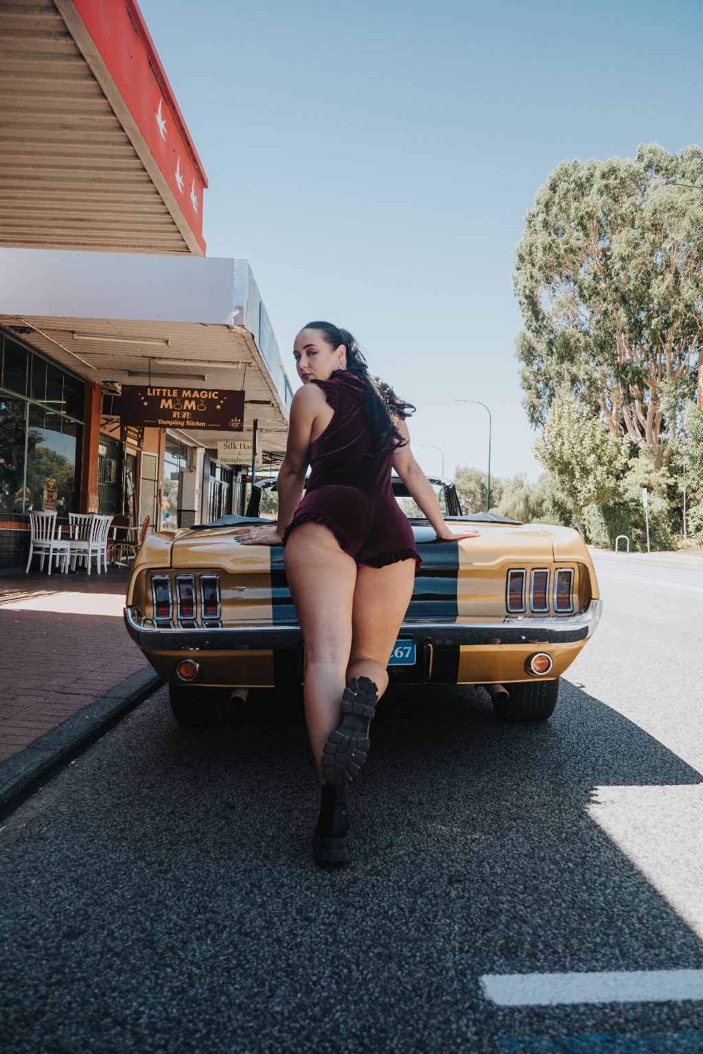 A person in a maroon bodysuit poses confidently on the hood of a classic yellow and black muscle car on a sunny street.