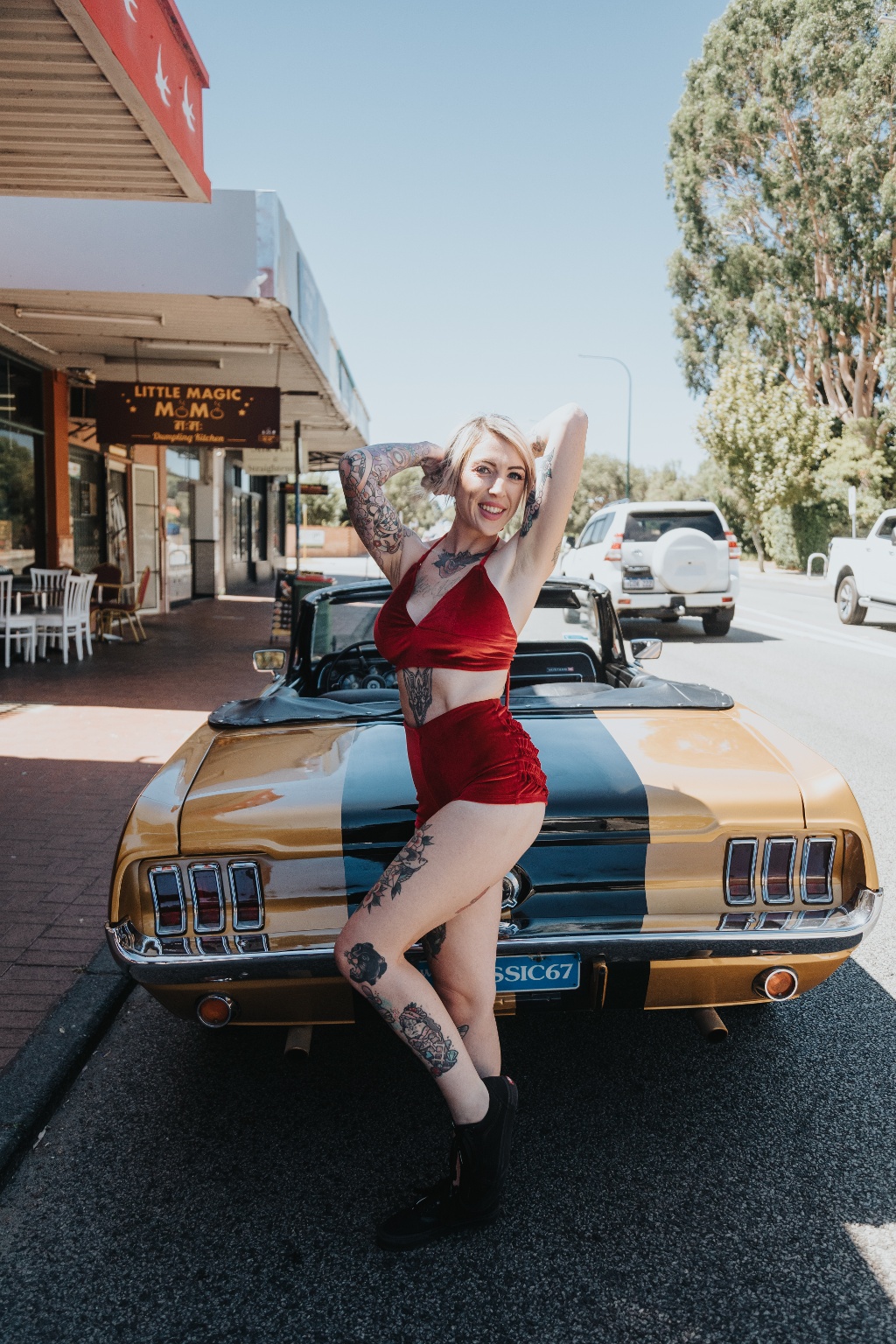 A person in a red swimsuit poses confidently on a vintage muscle car parked on a sunny street.
