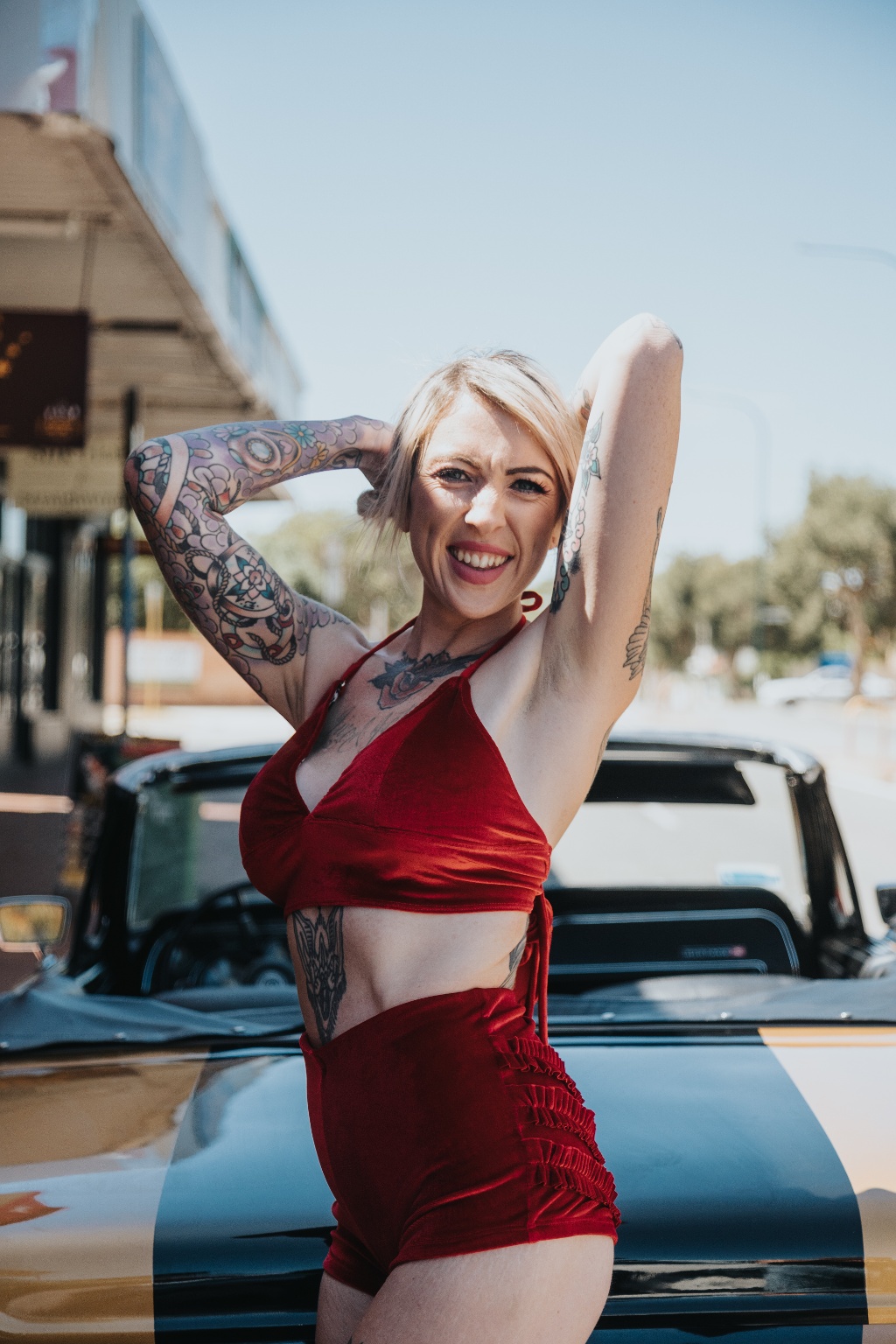 A person with colorful tattoos poses confidently in a red outfit against a classic car backdrop.