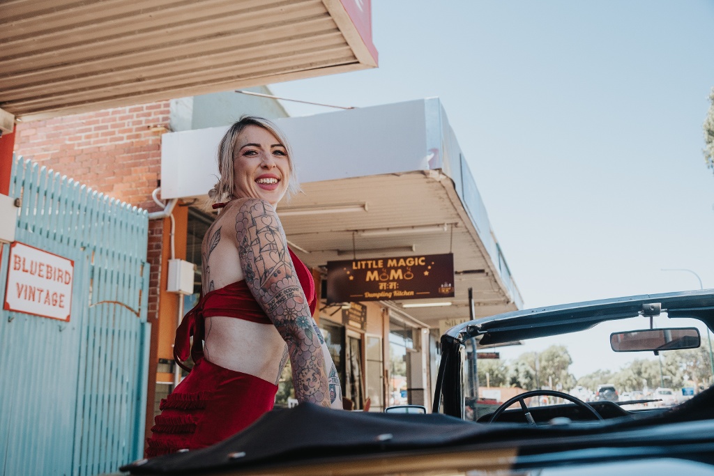 A person with tattoos on their arm is standing near a vintage car in front of a store.
