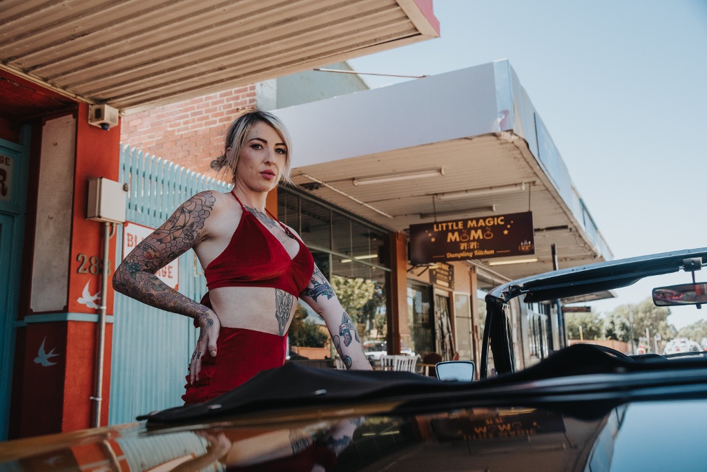 A person with tattoos stands confidently in front of a vintage car near a restaurant sign.
