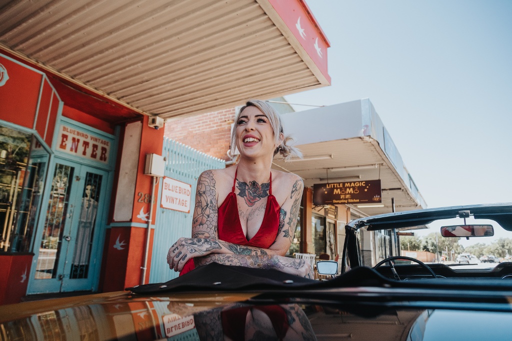 A person with tattoos and blonde hair leans on a car hood in front of a colorful storefront.